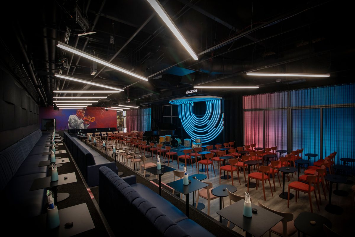 Dazzle main listening room with blue neon stage backdrop, intimate seating, and jazz mural