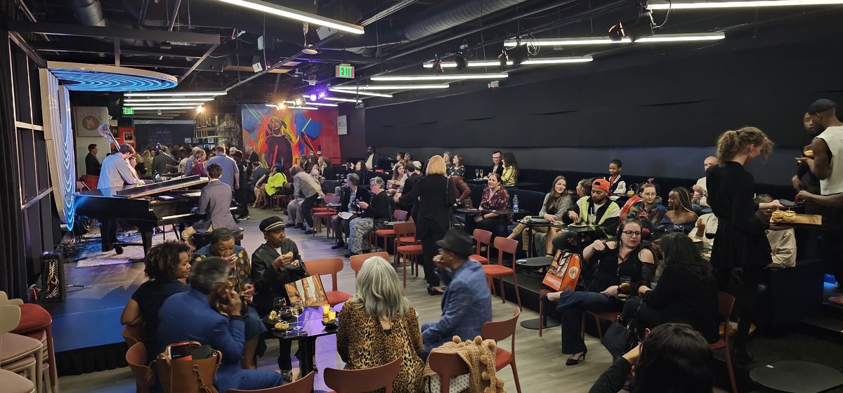 Packed audience at a Dazzle Denver event with live pianist and jazz mural in background